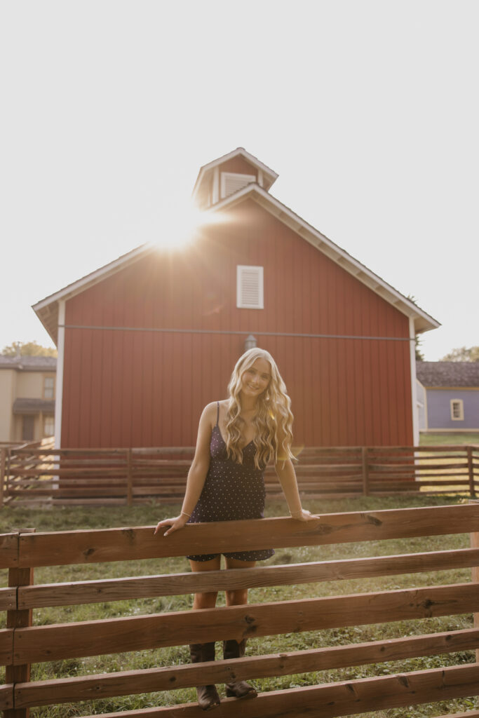 Senior girl leaning on a wooden fence with a red barn and sun flare behind her