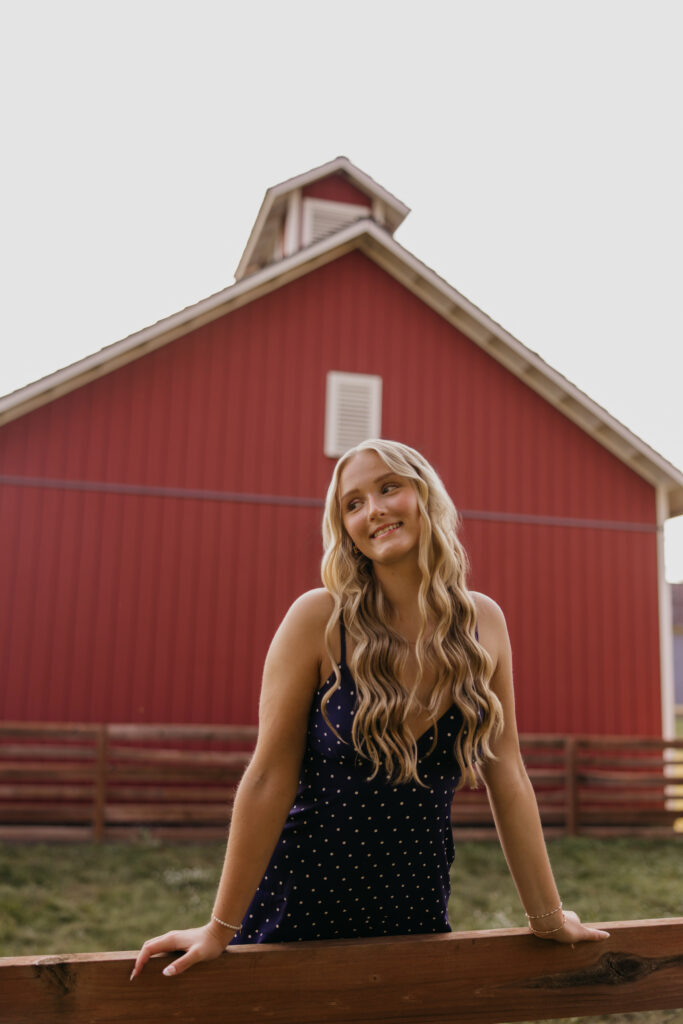 Senior girl leaning on a wooden fence rail in front of a red barn