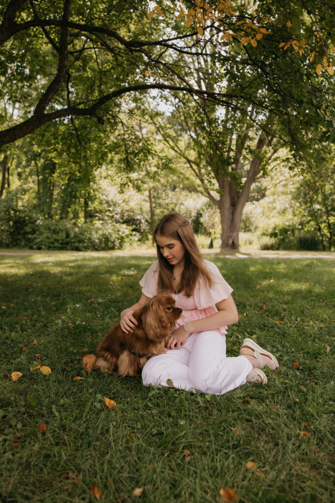 Senior girl kneeling on the grass petting her dog under a tree