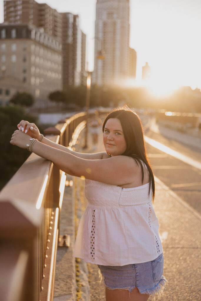 Senior girl leaning on a bridge railing with sun flare and skyline behind her