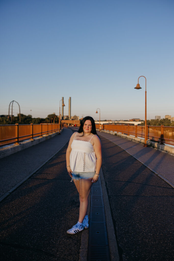 Senior girl in white top and denim shorts standing on a bridge at golden hour