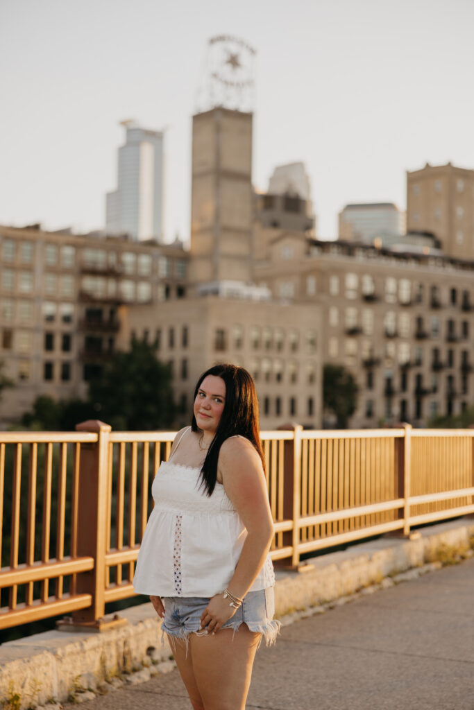Senior girl in white top and denim shorts smiling on a bridge with city skyline