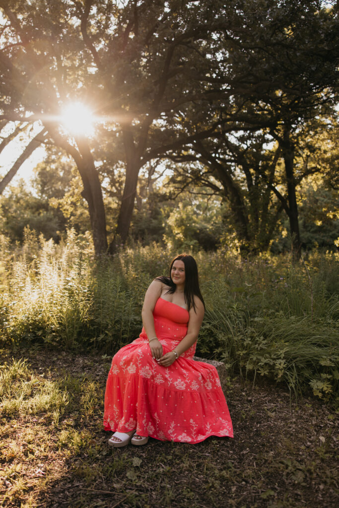 Senior girl in coral dress sitting under trees with golden sun flare behind her