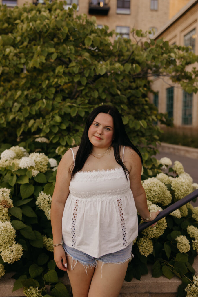 Senior girl in white top standing near white hydrangeas with a building behind her