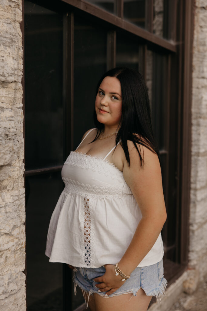 Senior girl in white eyelet top and denim shorts standing by a stone building