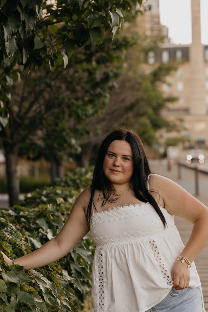 Senior girl in white top gesturing near greenery with a building in the background