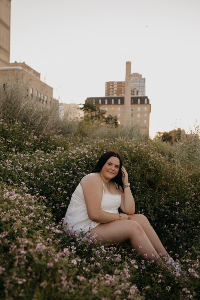 Senior girl in white top sitting among small wildflowers with skyline in the distance