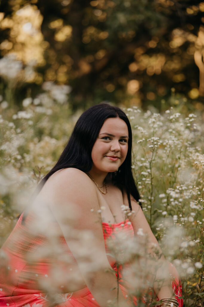 Senior girl in coral dress sitting among small white wildflowers