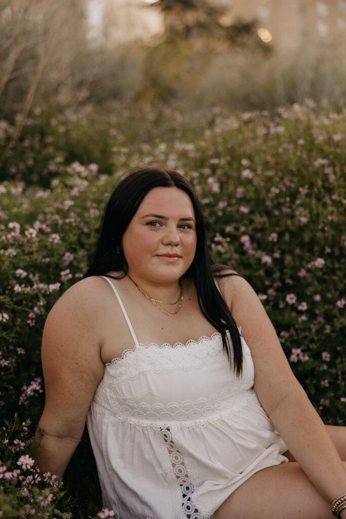 Senior girl in white top kneeling in a flower field near the Minneapolis skyline