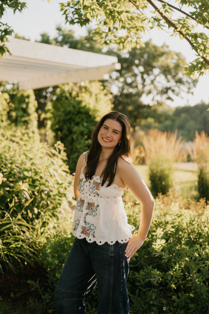 Senior standing near a garden pergola, wearing a white floral top and dark pants