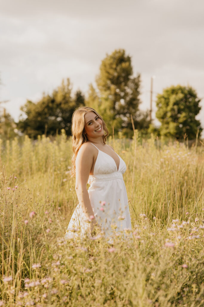 Senior girl standing in tall grass looking over her shoulder in golden hour light