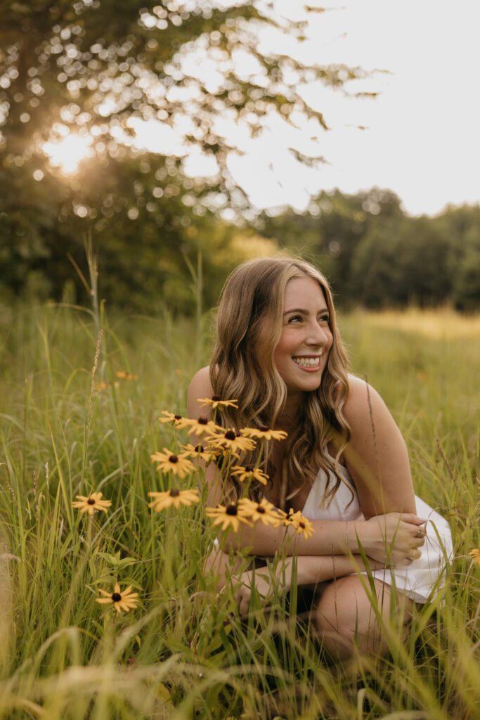 Senior girl crouching among wildflowers laughing in golden hour light in an open field