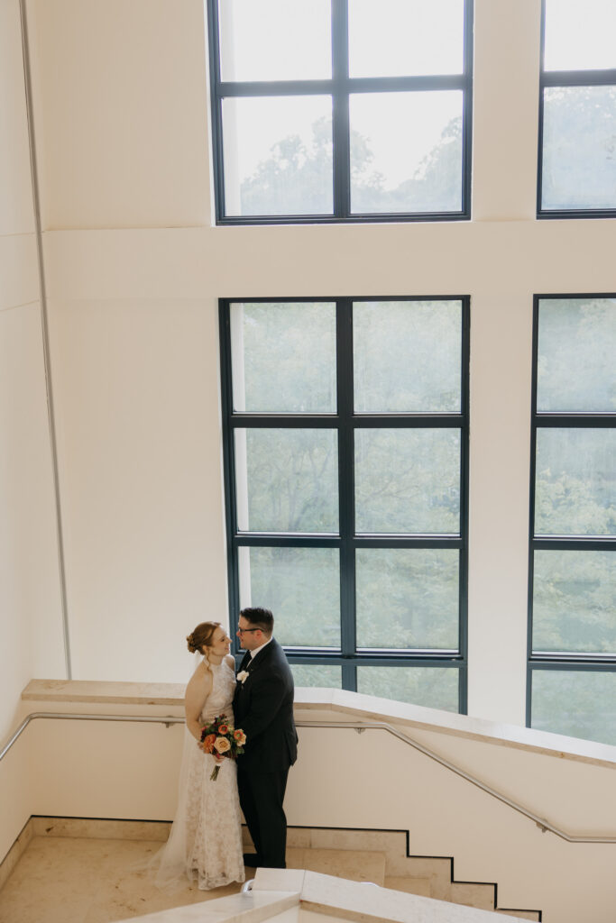 Wide shot of bride and groom on the staircase beneath large windows at Mia