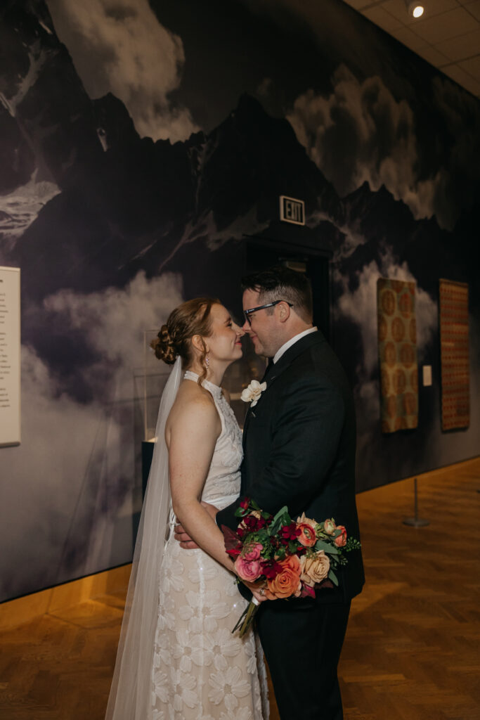 Bride and groom stand nose to nose in a dramatic mountain-mural gallery at their Minneapolis Institute of Art wedding, bride holding her bouquet