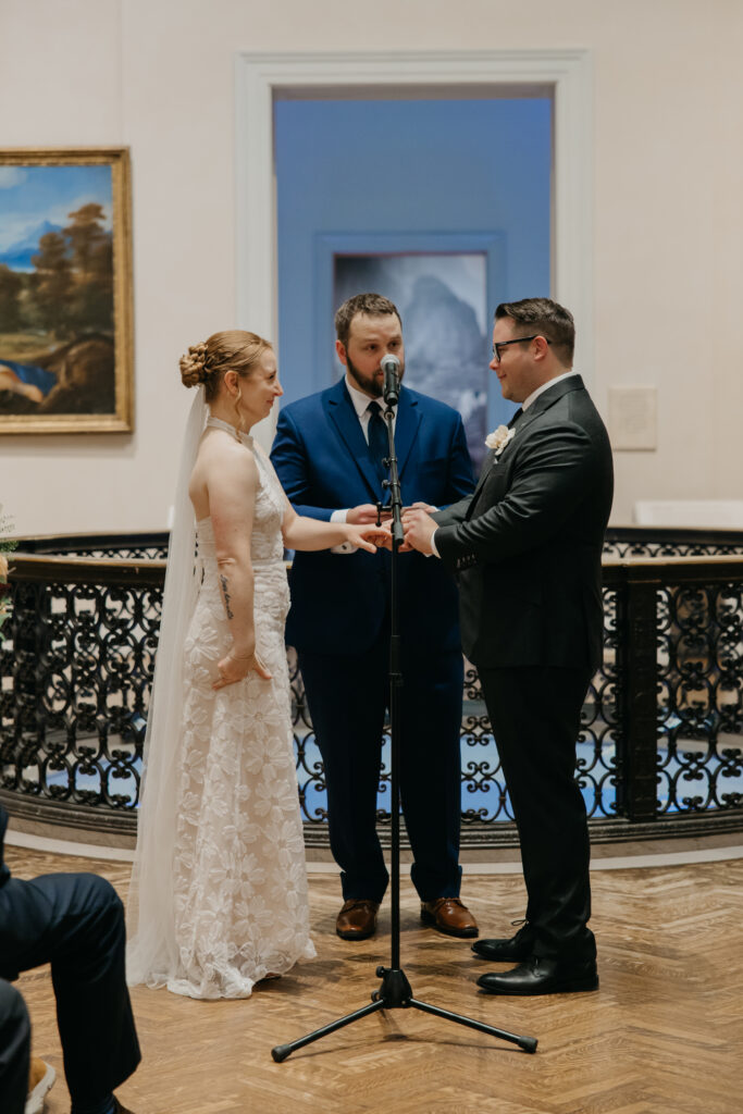 Bride and groom exchange rings at the microphone during their ceremony in the Baroque Gallery