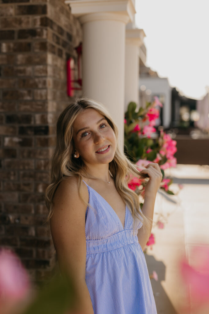 Eve beside a storefront window with pink flowers, in a blue striped dress