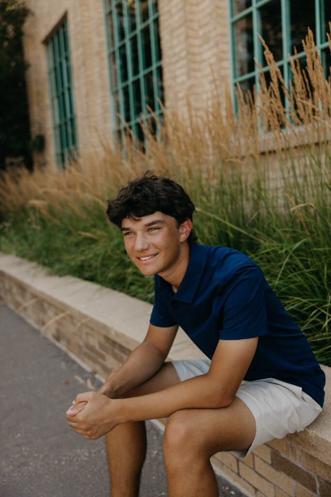 Senior in navy polo sitting on a stone ledge with tall grasses at Mill Ruins Park Minneapolis