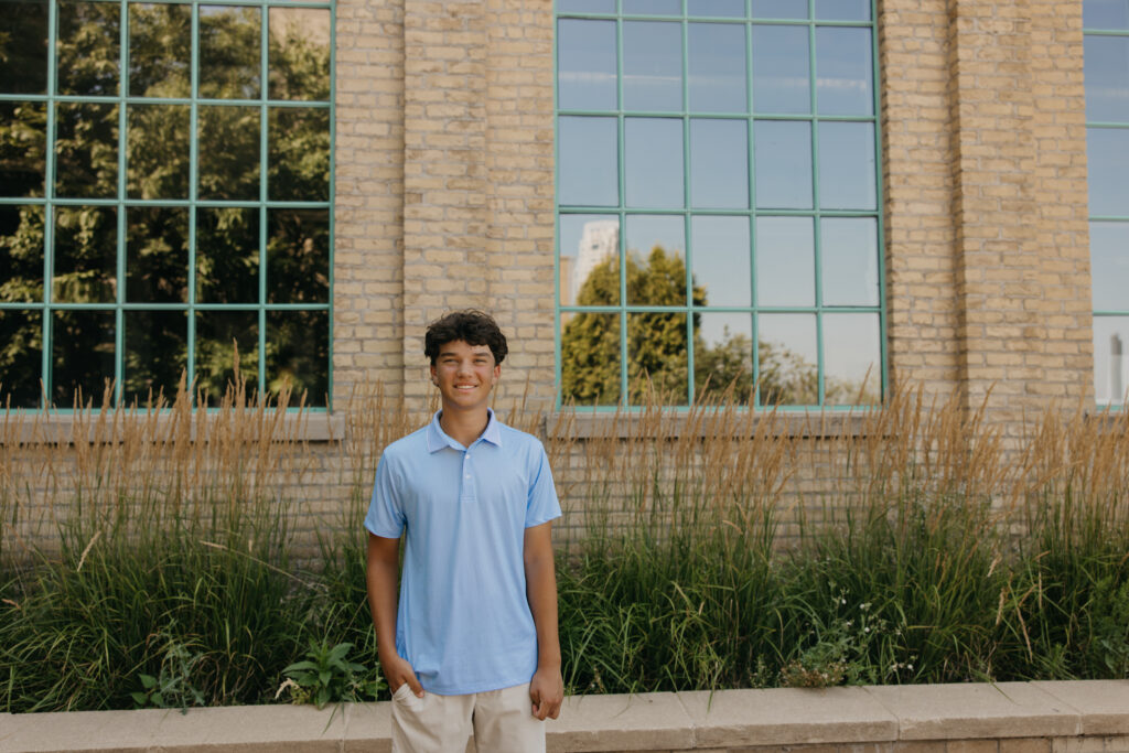 Senior in light blue polo standing near tall grasses with the brick building's windows reflected behind him