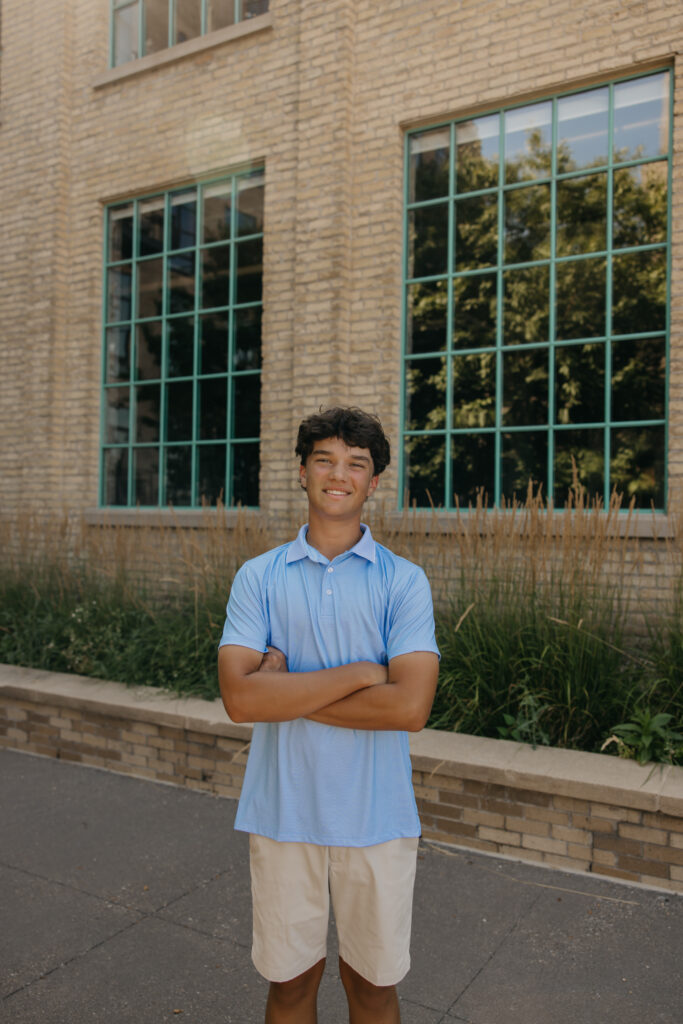 Senior in light blue polo standing arms crossed in front of a brick building with green-framed windows