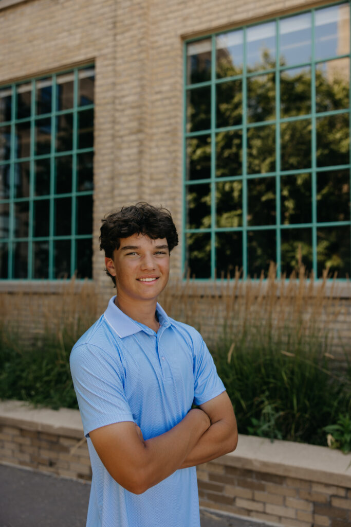 Senior in light blue polo standing arms crossed and smiling near the Mill Ruins Park brick building