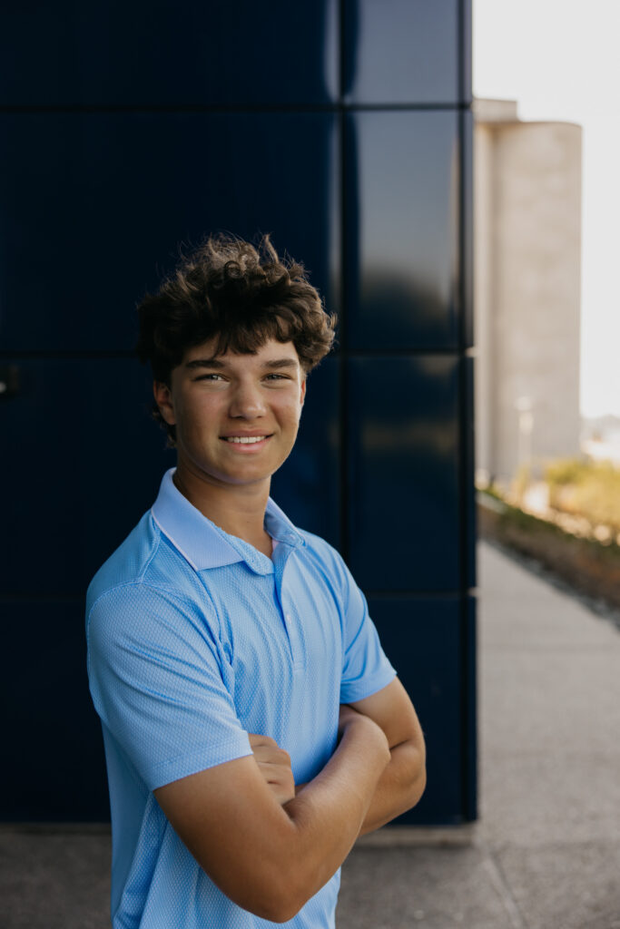 Senior in light blue polo standing arms crossed against a dark paneled building downtown Minneapolis