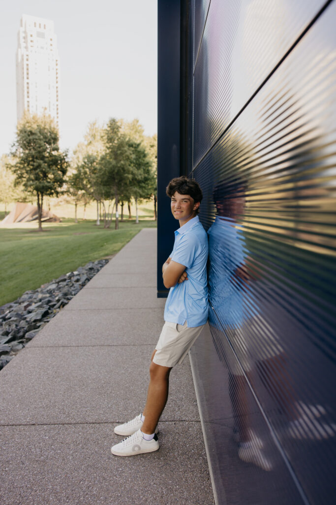 Senior in light blue polo leaning against a ribbed metal wall with the Minneapolis skyline behind him