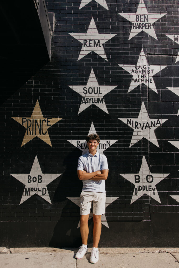 Senior in white polo standing arms crossed beneath the gold Prince star on the downtown Minneapolis mural