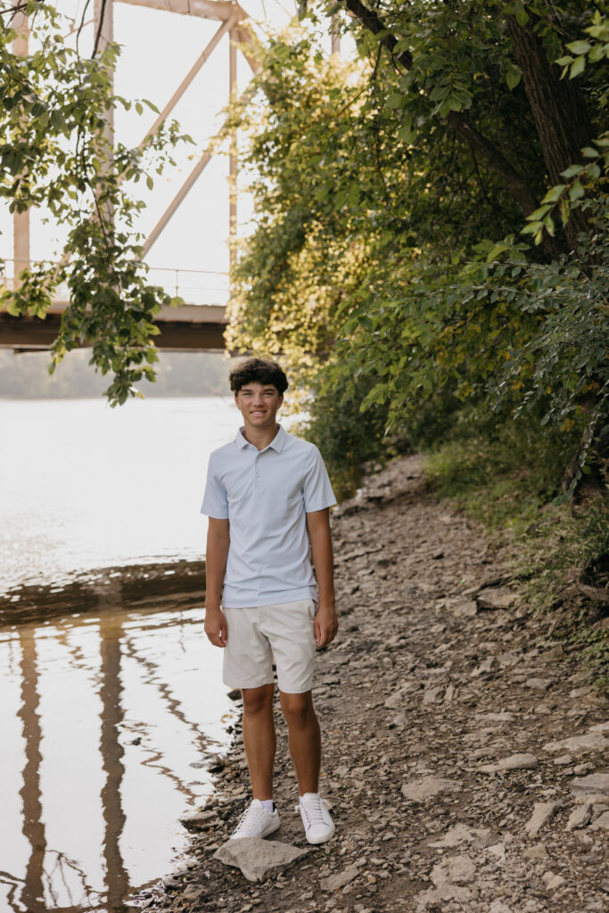 Senior in white polo standing at the water's edge with a rusted bridge behind him at Boom Island