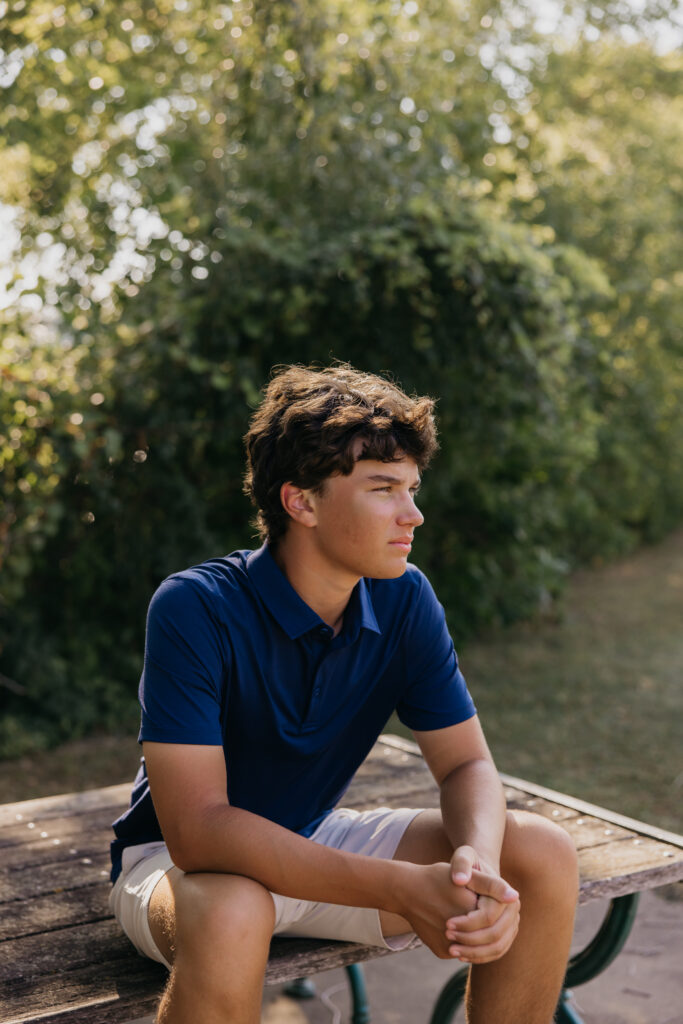 Senior in navy polo sitting in profile on a bench at Mill Ruins Park looking away from camera