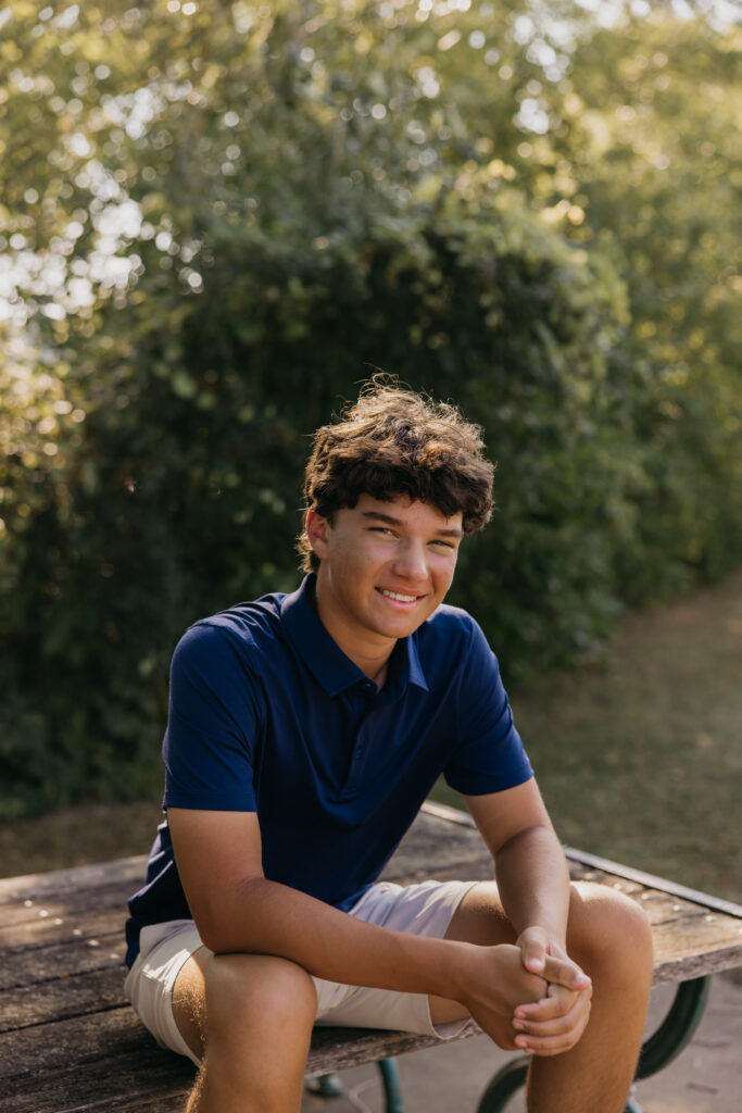 Senior in navy polo sitting on a wooden table at Mill Ruins Park, hands clasped, smiling