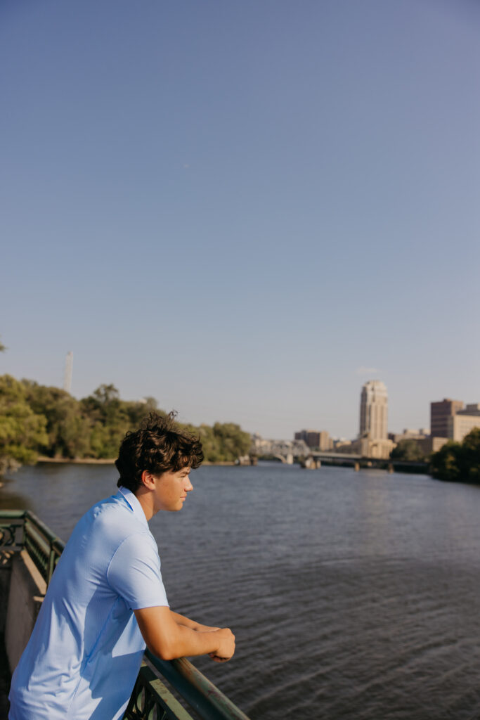 Senior in light blue polo leaning on a railing overlooking the river with the city skyline behind him