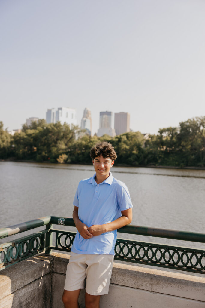 Senior in light blue polo standing at a railing with the Minneapolis skyline in the distance
