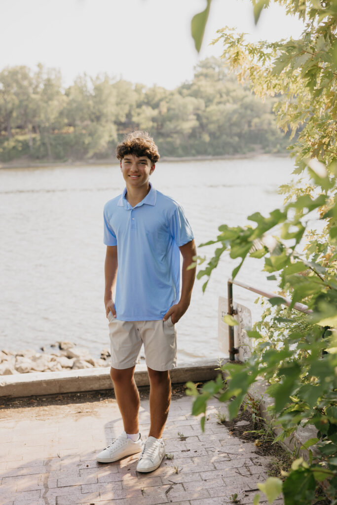 Senior in light blue polo standing on a brick path near the river at Boom Island Park