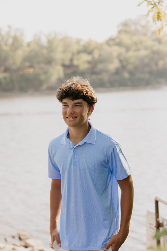 Senior in light blue polo smiling near the riverbank with trees in the background