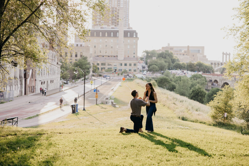 man kneeling to propose on a grassy hill with the Minneapolis skyline in the distance