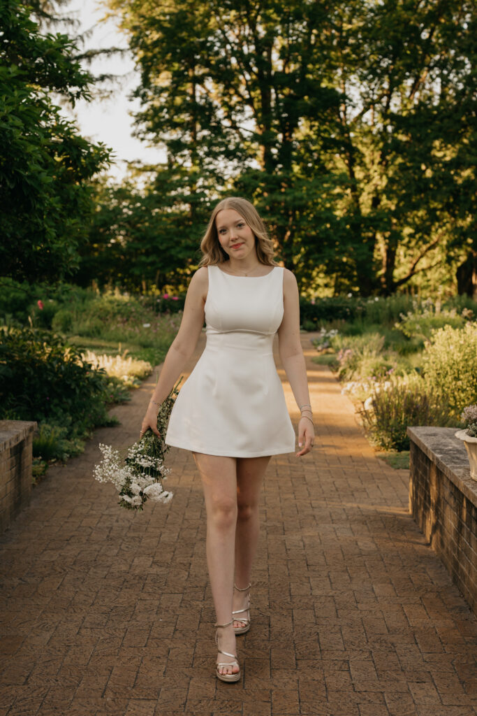 Senior girl in an ivory mini dress walking a garden path holding a small bouquet