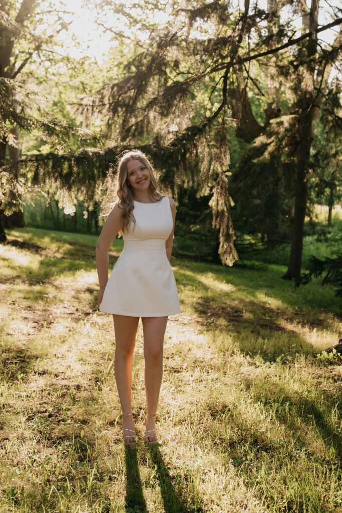 Senior girl in an ivory mini dress standing beneath a tree in dappled sunlight