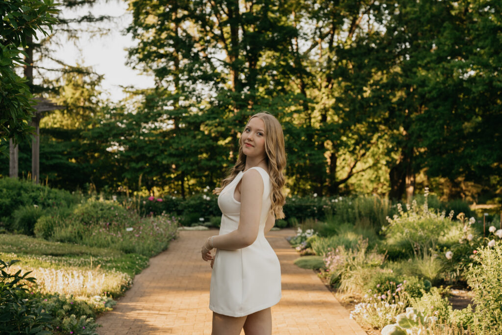 Senior girl in an ivory mini dress looking over her shoulder on a brick garden path