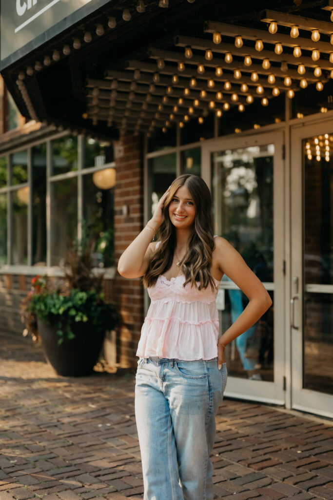 Senior girl in pink lace top and jeans under string lights near Aster Cafe at St. Anthony Main