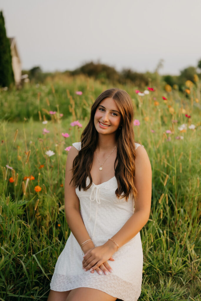 Senior girl in white dress kneeling in wildflower field with pink blooms at Pixel Dust Acres