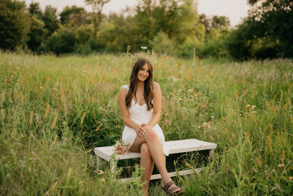 Senior girl in white dress sitting on white bench in field at Pixel Dust Acres