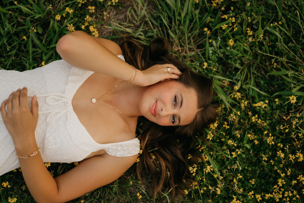 Senior girl in white dress lying in grass with wildflowers close to her face at Pixel Dust Acres