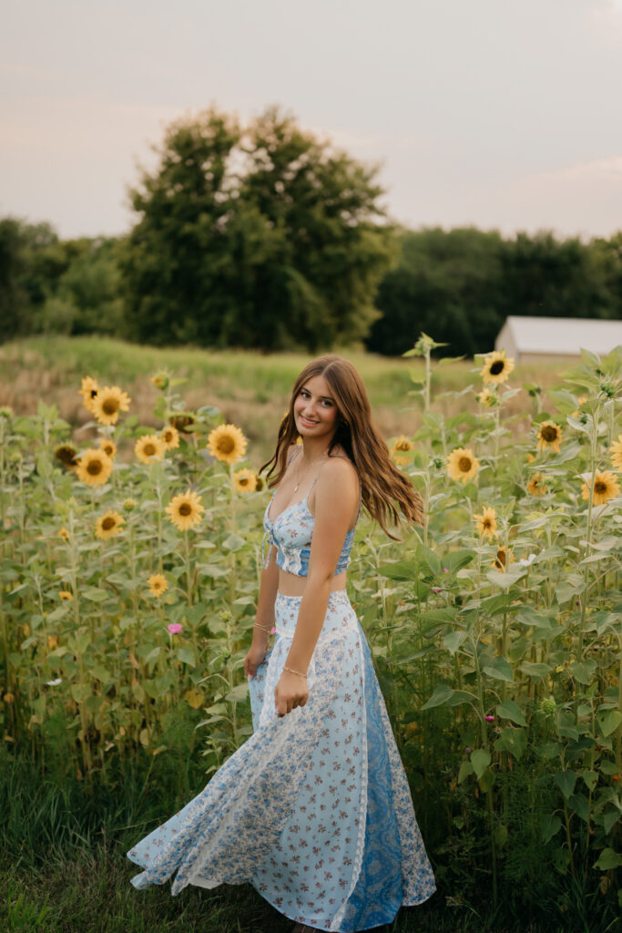 Senior girl in blue floral two-piece walking through sunflower field at Pixel Dust Acres