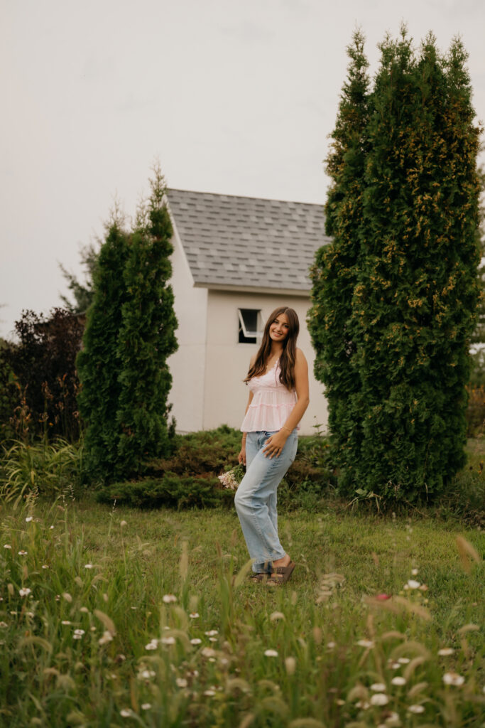 Senior girl in pink lace top standing near garden shed and evergreen trees at Pixel Dust Acres