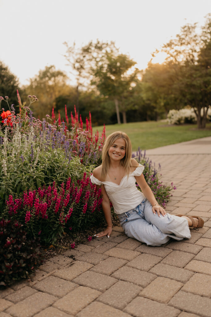 Senior sitting on a brick path near red and purple flowers, wearing a white top and jeans