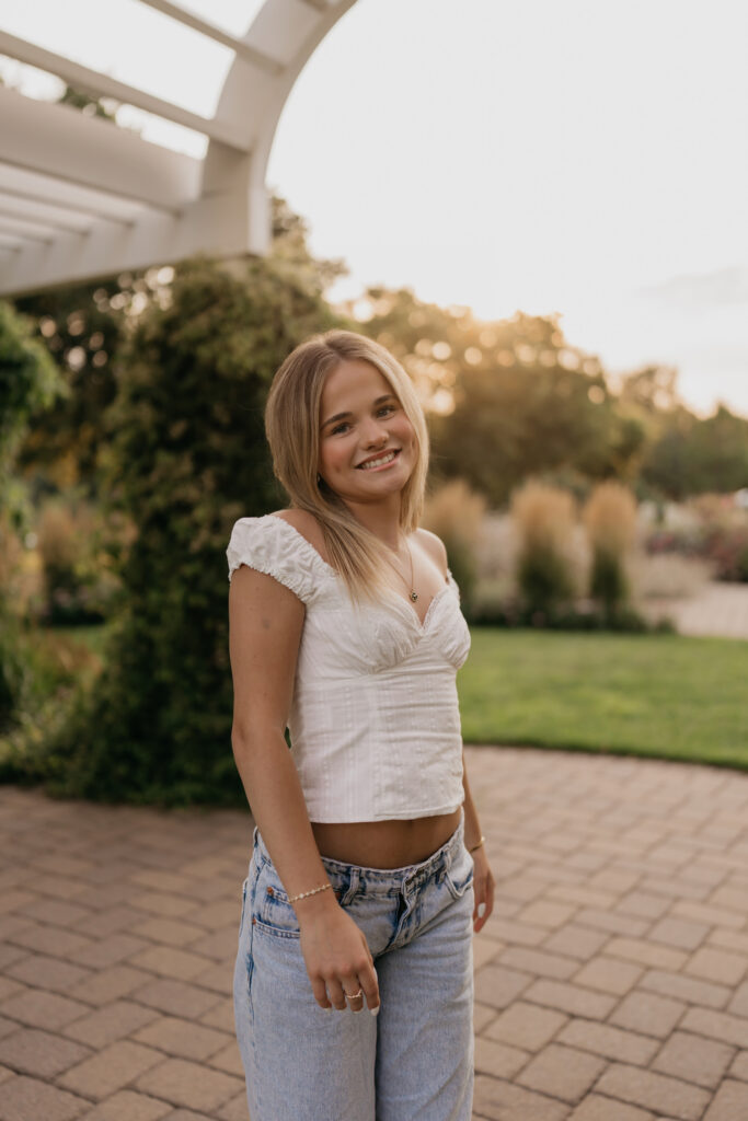 Senior standing on a brick path under a pergola, wearing a white top and jeans