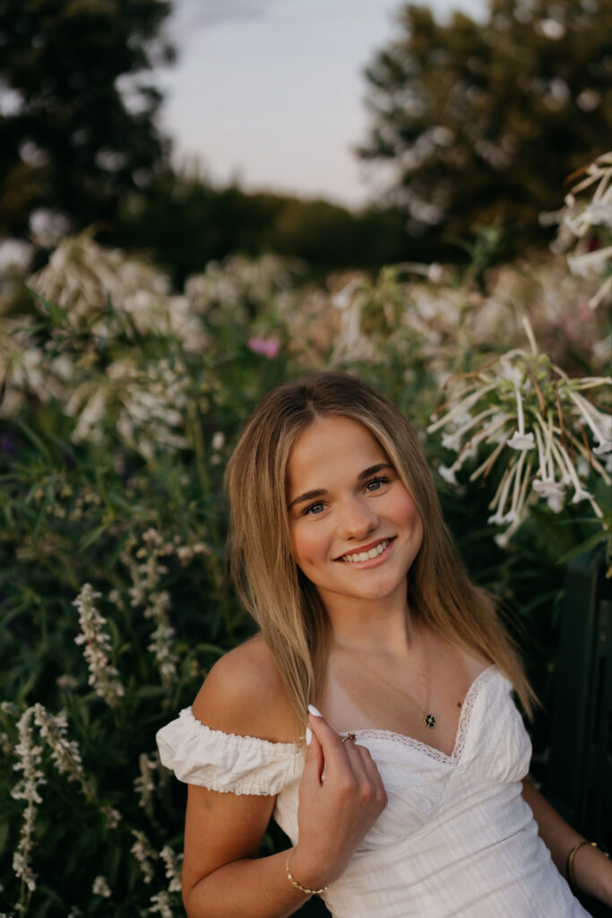 Senior smiling near white garden blooms, wearing an off-shoulder white top