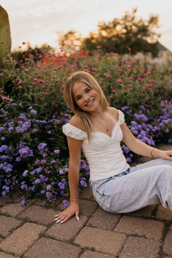 Senior sitting on a brick path surrounded by purple flowers, wearing a white top and jeans
