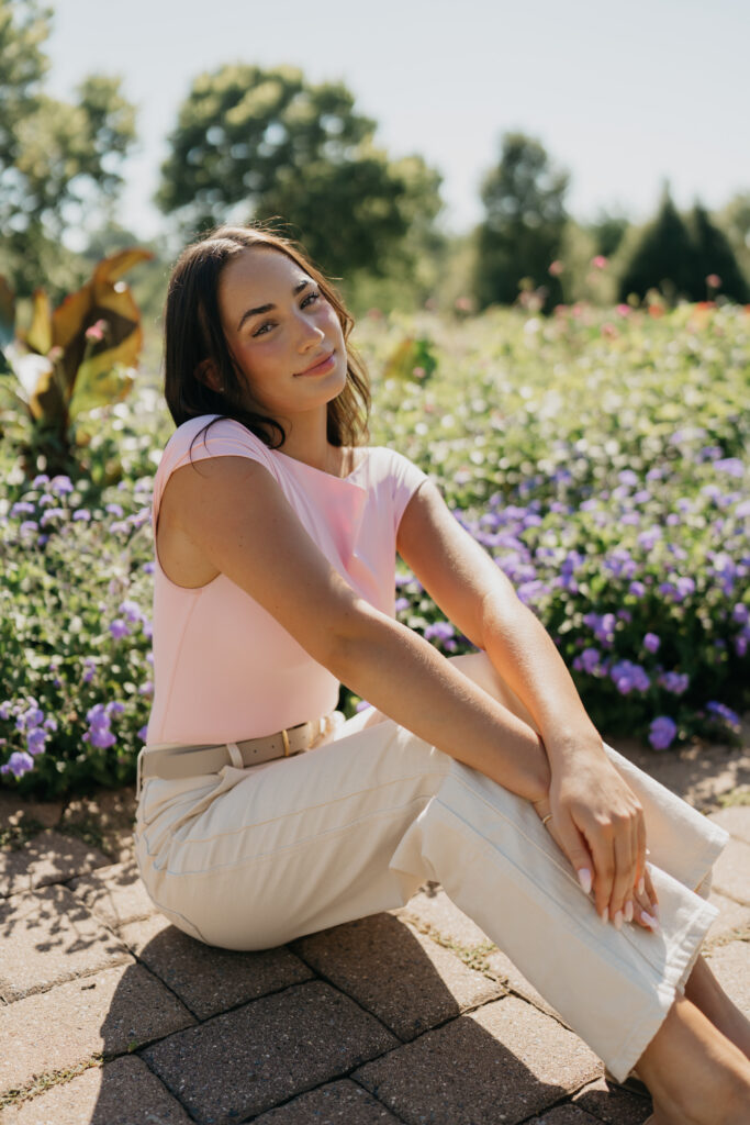 Senior girl in pink top and cream pants sitting on brick pavers in a garden