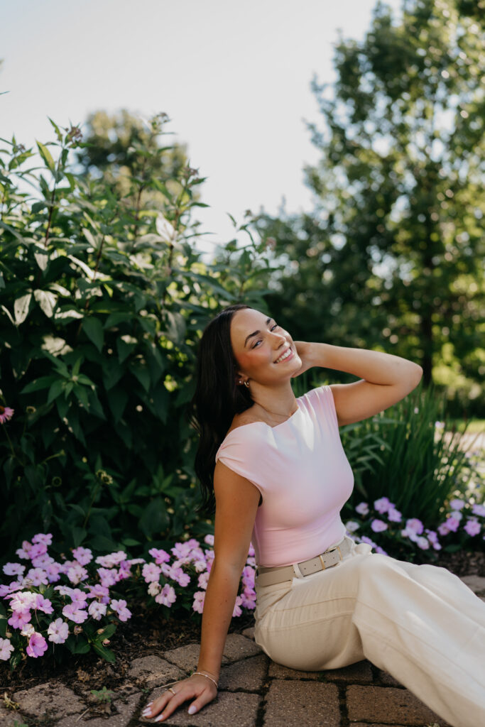 Senior girl in pink top leaning back on brick pavers surrounded by flowers